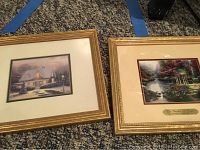 Two small framed Thomas Kinkade prints side by side on carpet with blue tape