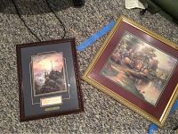 Two framed Thomas Kinkade prints on carpet, one with a gold-tone frame and cross image, the other with maroon matting.