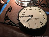 Double-sided Grand Central Station wall clock laid flat showing metal bracket and clock face with white background and black numbers.