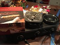 Photo showing the boxed Sur La Table Smores Grilling Kit next to the black Broul King HurryHot portable range with two coil burners and control knobs as well as part of the Tru triple slow cooker base.