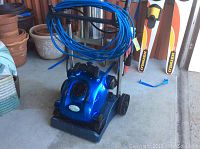 Blue robotic pool vacuum on floor with coiled cord and gardening pots visible
