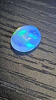 Oval opal gemstone on black textured surface showing blue body color with green and yellow flashes