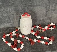 White ceramic Rae Dunn jar with red heart and lid beside red white pink bead garland