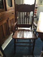 Front view of one wooden dining chair with armrests, spindle back, carved top back panel, and contoured solid wood seat in a medium brown finish.