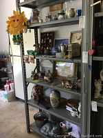 Five metal shelves holding assorted home decor items including candle holders, vases, decorative figurines, baskets, and a galvanized bucket. Items are placed for display in a garage setting.