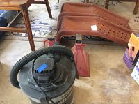Photo showing two red metal car ramps, a red jack stand, and a black and gray wet dry Shop-Vac vacuum cleaner with hose attached.