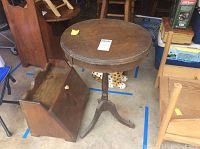 View of the round wooden table standing next to the wooden bread box on the floor with various other wooden furniture items visible.