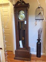 Full view of the tall wooden grandfather clock showing carved details on top, glass front door, and face with Roman numerals.