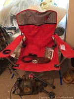 Red UGA folding chair with grey accents, armrests and cup holder on left arm, sitting on carpeted floor. Brown leather belt and leopard print flask on chair seat. Crown Royal bottle displayed near chair base seen.