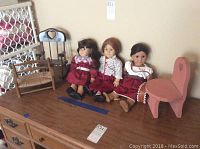Three American Girl dolls seated on a wooden dresser alongside two pieces of doll furniture, including a rocking chair and a pink chair with a heart cut-out.