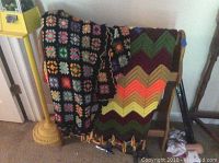 Wooden quilt rack holding several crocheted blankets showing multicolored granny square pattern and zigzag striped pattern.