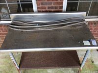 Front view of stainless steel table top with rusted lower shelf and metal legs with rust. Table has a backsplash panel at the back and a hose resting on top, hose not included.