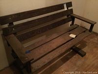 Front view of the wooden bench placed indoors against a wall, showing slatted wood construction and armrests.