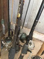 Photo showing several fishing rods with reels attached standing upright in a garage corner, displaying black handles and various reel types.