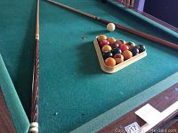 Full pool table with balls arranged in triangle rack and two pool cues on table surface.