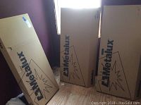 Three large cardboard boxes labeled Metalux standing upright in a room with purple walls and wooden floor, showing packaging of ceiling lights.