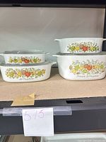 Five CorningWare La Marjolaine casserole dishes with lids stacked on shelf