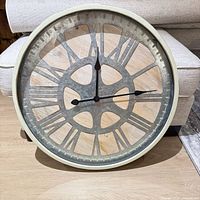 Front view of 24-inch rustic wall clock showing cream frame, glass front, Roman numeral cutouts, wood-look background