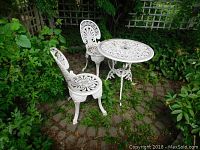 Wide view of white cast aluminum patio table and two chairs on brick patio surrounded by greenery.