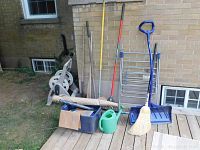 Photo showing garden tools arranged against a wall including hose rack, axe, rake, watering can, broom, cardboard box, and plastic container.
