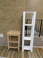 Wood kitchen island cart and white shelving unit placed upright on wooden deck against brick wall and black railing.
