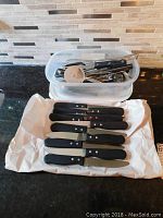 Photo shows six black-handled steak knives laid out on white paper. Knives have three rivets each and serrated blades.