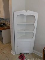 Full front view of white painted wooden corner cabinet showing three open shelves and lower closed cabinet door.