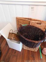 View of five woven baskets of various shapes and sizes stacked and displayed together, the largest basket is dark brown and round with handles, two medium rectangular natural tone baskets with cutout handles, a small narrow dark brown basket, and an elongated natural tone basket with tall handles
