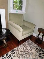 Full view of the fabric upholstered easy chair with armrests and wooden legs positioned near a window and side tables.