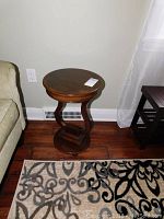 Full view of the round wooden side table placed on a wooden floor beside a couch and near a wall outlet, showing its shape and base design.