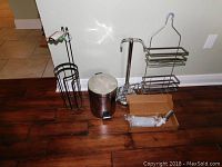 Full view of lot showing black metal toilet paper holder, small stainless steel step trash can, silver metal shower rack, white soap dispenser with pump, and wall-mounted silver metal toilet paper holder.