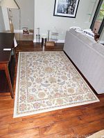 Full view of the carpet positioned on hardwood floor next to a gray sofa and dark wood furniture, showing the floral border and central design.