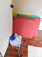 Photo showing multiple interlocking rubber floor tiles in red, yellow, green, and blue colors stacked against the wall next to a humidifier and heating pad on the floor.