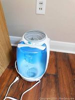 Front angle view showing white base with blue water tank and power cord laid out on wooden floor.