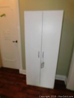 White faux wood cabinet closed, showing two doors with silver handles, standing upright on hardwood floor next to wall and door.