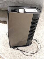 Side view of the black Oreck air purifier showing its slim rectangular body, the attached power cord, and the small attached cleaning brush on top.