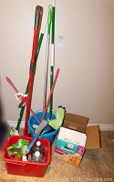 Photo of various mops with handles standing upright, blue and green plastic buckets, red bin filled with assorted cleaning chemicals and bottles, and a cardboard box nearby.