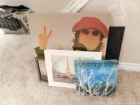 Full view of the collection of five artworks leaning against wall on carpeted floor, showing portrait, Eiffel Tower print, and other pieces.