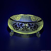 Bowl glowing bright yellow-green under UV light highlighting uranium content, shows black scrollwork and tripod feet