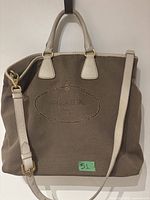 Front view showing canvas body, leather handles, detachable strap, gold hardware and embroidered crest