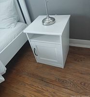 Front angled view of white nightstand next to bed showing cabinet door and handle
