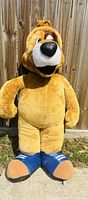 Full view of the five-foot tan dog plush standing against wooden fence