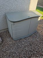 Front view of beige/light gray plastic outdoor storage box placed on gravel