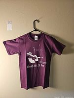 Full view of burgundy KKUP event t-shirt on hanger