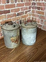 Front view of both galvanized water cans on wood surface against brick wall