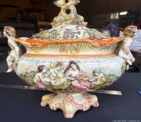 Front view of porcelain tureen showing relief figures, cherub handles, pedestal base, lid with finial