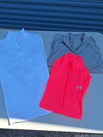 Overall shot of three long-sleeve athletic tops laid out: white, neon pink Under Armour, grey