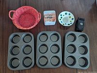 All items arranged on table: three muffin pans, red wicker basket, graphic tin, enamelled dish, magnetic wrist strap