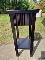 Full front view showing tall narrow black side table with drawer and lower shelf