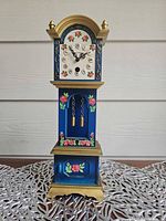 Full frontal view of miniature grandfather clock showing blue body, floral painting, gold trim